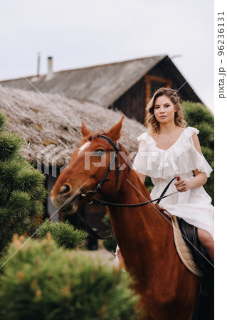 A woman in a white sundress riding a horse near a farm A woman in a white sundress riding a horse near a farm 96236131