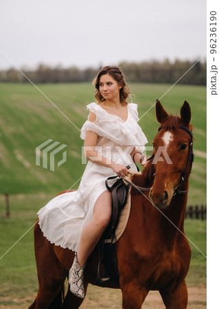 A woman in a white sundress riding a horse in a field A woman in a white sundress riding a horse in a field 96236190