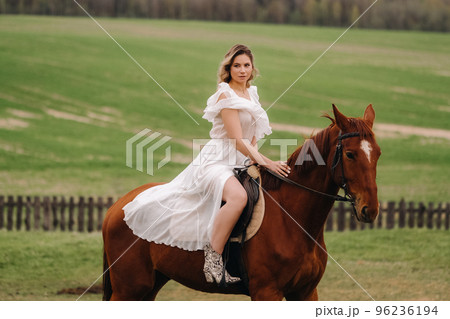 A woman in a white sundress riding a horse in a field A woman in a white sundress riding a horse in a field 96236194