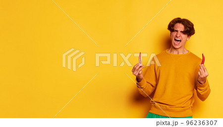 Portrait of young man with curly hair emotively posing with chili pepper isolated over yellow background. Win. Flyer 96236307