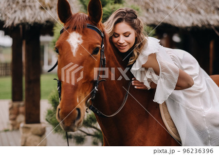 A woman in a white sundress riding a horse near a farm 96236396