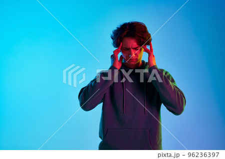 Portrait of young man with curly hair posing with hands to temples isolated over blue background in neon light. Thinking 96236397