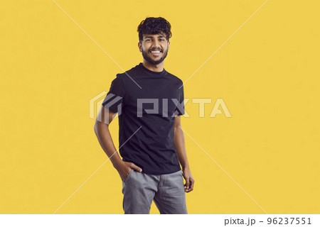 Young South Asian man with dark curly hair posing with smile in casual clothes stands in studio 96237551