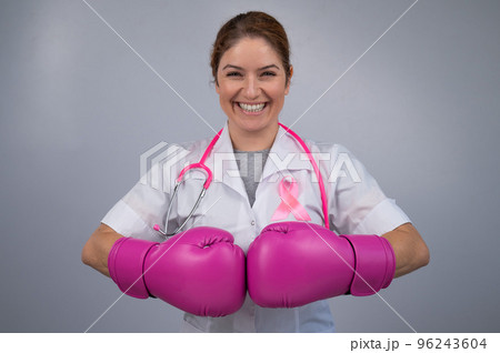 Smiling female doctor in pink boxing gloves with a pink ribbon on a gray background. Fight against breast cancer. Smiling female doctor in pink boxing gloves with a pink ribbon on a gray background. Fight against breast cancer. 96243604