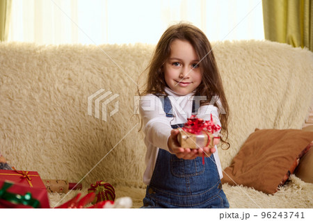 Portrait of beautiful European little girl dressed in white pull over and denim overalls, cutely smiles while handing to camera a Christmas gift, sitting on a comfortable sofa in a cozy home interior. 96243741
