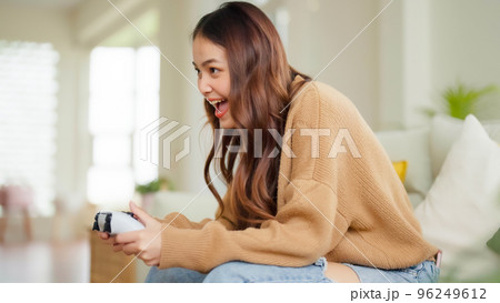 Beautiful young asian woman sitting in living room sofa holding joystick Beautiful young asian woman sitting in living room sofa holding joystick 96249612