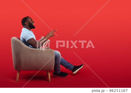 Amused televiewer sitting in the armchair at home while eating popcorns and watching a movie on red background Amused televiewer sitting in the armchair at home while eating popcorns and watching a movie on red background 96251167