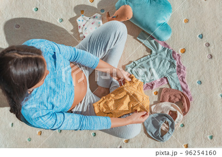 Happy pregnant woman is sitting with presents and at baby shower. Pregnancy shopping for baby or gift concept with woman looking at baby clothing bodysuits and bibs Happy pregnant woman is sitting with presents and at baby shower. Pregnancy shopping for baby or gift concept with woman looking at baby clothing bodysuits and bibs 96254160