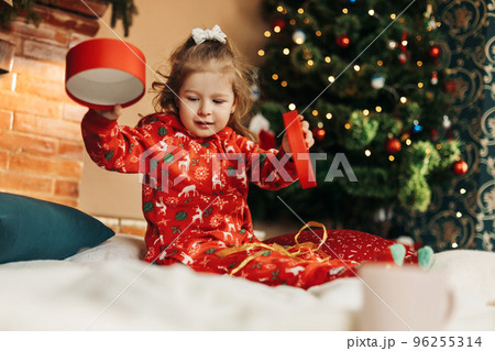 A little girl opens a Christmas present near the Christmas tree. Christmas time, gifts, festive mood. The concept of Christmas and New Year 96255314