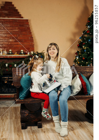 Cute mom and daughter are looking through a family photo album on Christmas Eve, spending time together by the fireplace and the Christmas tree. Christmas, New Year, memories, family evening 96255857