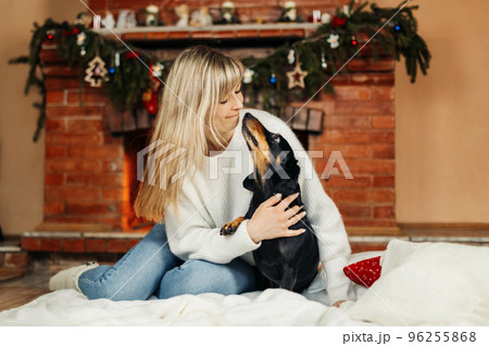 A smiling young woman is playing with a dog, sitting on the floor near the fireplace, on Christmas Eve. Christmas mood, Christmas holidays. The concept of Christmas and New Year A smiling young woman is playing with a dog, sitting on the floor near the fireplace, on Christmas Eve. Christmas mood, Christmas holidays. The concept of Christmas and New Year 96255868