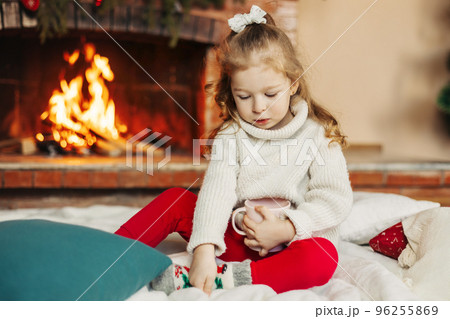 A baby girl drinks hot chocolate by the fireplace at home. A little girl on Christmas Eve or New Year's Eve at home. The concept of Christmas and New Year A baby girl drinks hot chocolate by the fireplace at home. A little girl on Christmas Eve or New Year's Eve at home. The concept of Christmas and New Year 96255869