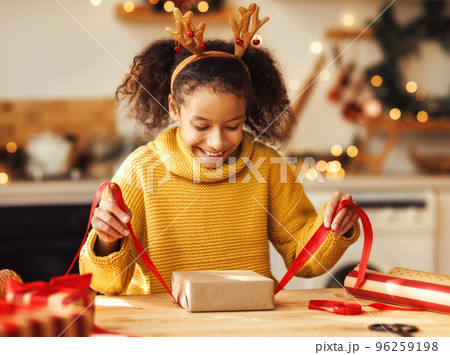 Cheerful ethnic girl decorating gift box on Christmas day Cheerful ethnic girl decorating gift box on Christmas day 96259198