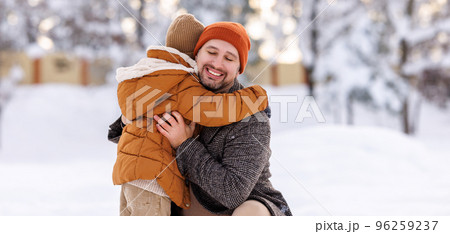 Happy family enjoying snowy weather outdoor, dad with little son hugging during walk in winter park 96259237