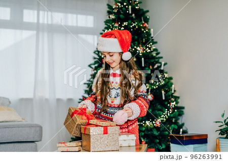 A happy smiling little girl in Santa hat and mom's festive knitted sweater packing gift boxes on the Christmas tree background. Christmas mood concept. Preparing or opening presents. Selective focus. A happy smiling little girl in Santa hat and mom's festive knitted sweater packing gift boxes on the Christmas tree background. Christmas mood concept. Preparing or opening presents. Selective focus. 96263991