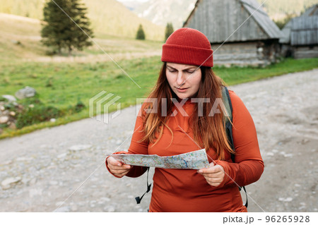 Sad female tourist is exploring new locations on mountains background. Redhead girl holding a paper map in a sunny day. Travel concept. Hiking woman wearing knit hat exercising outdoor Sad female tourist is exploring new locations on mountains background. Redhead girl holding a paper map in a sunny day. Travel concept. Hiking woman wearing knit hat exercising outdoor 96265928