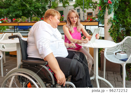 Mother with disability in wheelchair and her daughter using digital tablet while sitting at the table in cafe Mother with disability in wheelchair and her daughter using digital tablet while sitting at the table in cafe 96272252