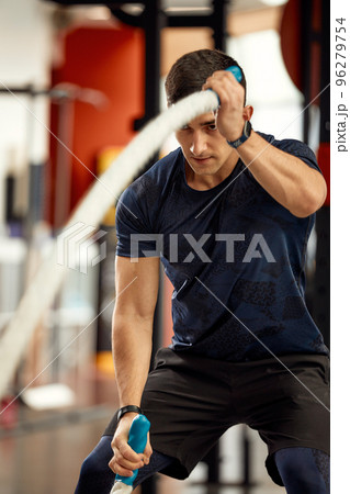 young handsome man in a black t-shirt is engaged with ropes in the gym on a window background. young handsome man in a black t-shirt is engaged with ropes in the gym on a window background. 96279754