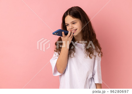 Mobile smart voice technology. Portrait of little girl wearing white T-shirt talking to smartphone using virtual assistant, digital speaker app. Indoor studio shot isolated on pink background. Mobile smart voice technology. Portrait of little girl wearing white T-shirt talking to smartphone using virtual assistant, digital speaker app. Indoor studio shot isolated on pink background. 96280697