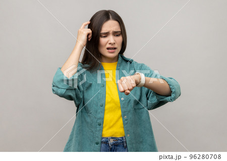 Portrait of confused puzzled woman pointing at her watch, hurry up, deadline, worrying about delay, wearing casual style jacket. Indoor studio shot isolated on gray background. 96280708