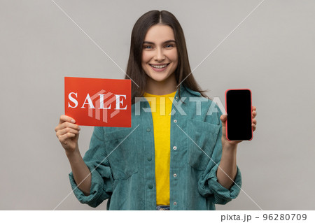 Cheerful woman holding sale inscription, cellphone and smiling at camera, showing discounts on mobile device, application, wearing casual style jacket. Indoor studio shot isolated on gray background. 96280709