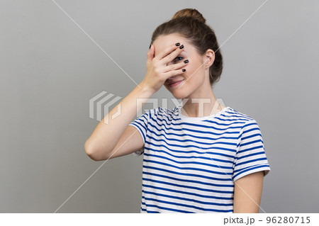 Portrait of interested woman wearing striped T-shirt covering eyes with hand, peeking through fingers with happy curious expression. Indoor studio shot isolated on gray background. Portrait of interested woman wearing striped T-shirt covering eyes with hand, peeking through fingers with happy curious expression. Indoor studio shot isolated on gray background. 96280715