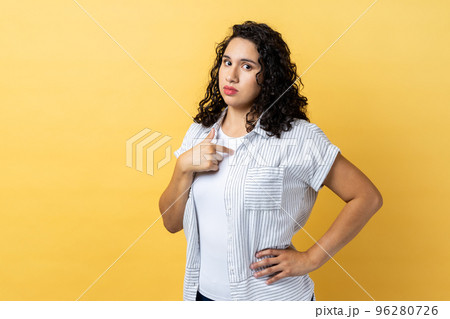 Portrait of confident woman with dark wavy hair pointing at herself, feeling proud and self-important, keeps hand on hip, having big ego. Indoor studio shot isolated on yellow background. 96280726