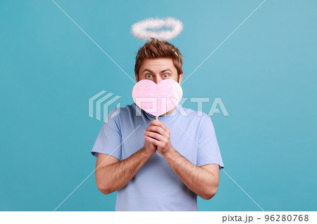 Portrait of angelic man with holy nimbus covering half of face with pink paper heart on stick, being shy to demonstrate love care, romantic feelings. Indoor studio shot isolated on blue background. 96280768