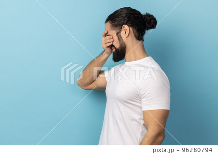 Don't want to look at this. Side view of scared man with beard wearing white T-shirt standing, covering eyes with hand, refusing to watch. Indoor studio shot isolated on blue background. 96280794