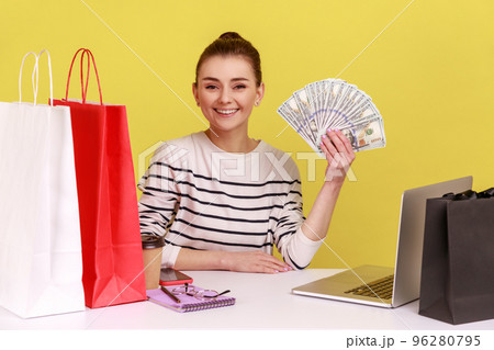 Portrait of satisfied woman office manager sitting on workplace with shopping bags, showing fan of dollars, cashback, looking at camera. Indoor studio studio shot isolated on yellow background. 96280795