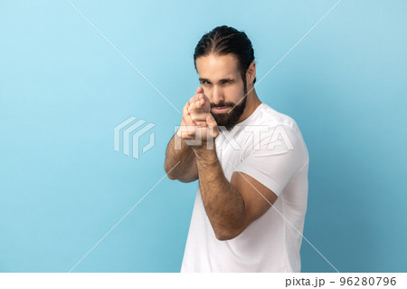 Portrait of assertive dangerous man with beard wearing white T-shirt pointing fingers at target imitating weapon making shot, terrorism. Indoor studio shot isolated on blue background. 96280796