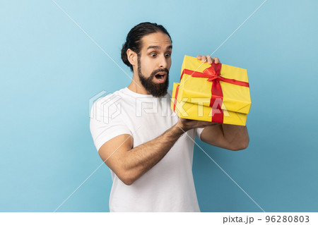Portrait of handsome man with beard wearing white T-shirt unpacking present, looking inside box with shocked and scared expression, birthday gift. Indoor studio shot isolated on blue background. 96280803