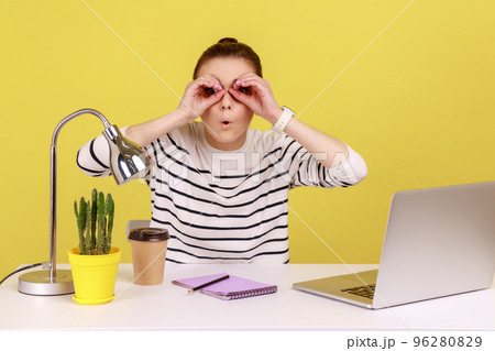 Astonished shocked woman office worker sitting at workplace holding hands near eyes pretending to look in binoculars, find something surprised. Indoor studio studio shot isolated on yellow background. Astonished shocked woman office worker sitting at workplace holding hands near eyes pretending to look in binoculars, find something surprised. Indoor studio studio shot isolated on yellow background. 96280829