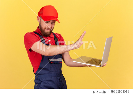 Side view portrait of handsome worker man wearing blue uniform and red cap holding notebook, forbidden information, showing stop ban gesture. Indoor studio shot isolated on yellow background. Side view portrait of handsome worker man wearing blue uniform and red cap holding notebook, forbidden information, showing stop ban gesture. Indoor studio shot isolated on yellow background. 96280845