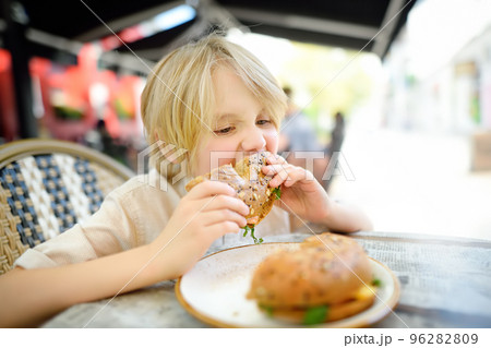 Cute blonde boy eating large bagel with salmon and arugula in outdoor fast food restaurant 96282809