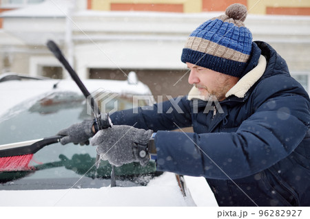 Mature man cleaning car using brush after snowfall. Snowdrift after snowstorm on the hood of automobile. Difficult transport situation on roads in city after snow storm. Winter weather. 96282927