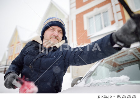 Mature man cleaning car with brush after snowfall. Snowdrift after snowstorm on the hood of automobile. Difficult transport situation on roads in city after snow storm. Winter weather. Mature man cleaning car with brush after snowfall. Snowdrift after snowstorm on the hood of automobile. Difficult transport situation on roads in city after snow storm. Winter weather. 96282929