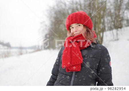 Close-up portrait of young beautiful caucasian woman with pink hair wearing red hat and scarf during snowfall on winter day. 96282954