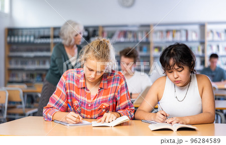 Two teenage friends sit at a desk in school library and carefully do assignments 96284589