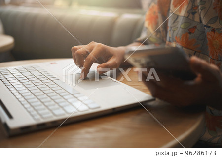 Close-up of woman's hand using smartphone while working on laptop in a coffee shop. 96286173