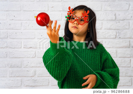 Asian girl in masquerade glasses with reindeer horns, playing with a Christmas ball Asian girl in masquerade glasses with reindeer horns, playing with a Christmas ball 96286688