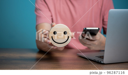 Man using Mobile Phone and holding wooden disc with smiling face icons, customer satisfaction service, feedback, rating and positive customer review, experience, world mental health day, think 96287116