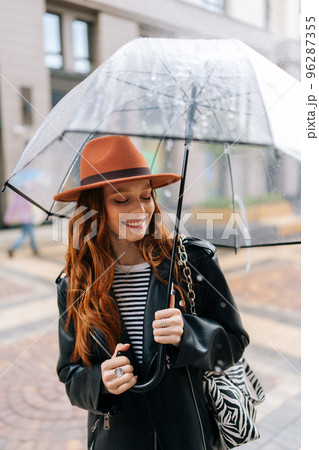 Vertical shot of cheerful young woman in hat standing with transparent umbrella on city street enjoying rainy weather outdoors, smiling looking down. Concept of female lifestyle at autumn season. 96287355