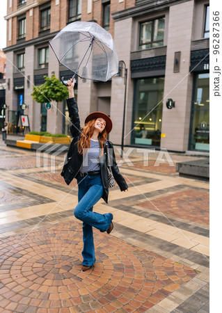 Vertical full length portrait of cheerful young woman in hat dancing and having fun with transparent umbrella on beautiful city street, enjoying rainy weather outdoors. Concept of female lifestyle. Vertical full length portrait of cheerful young woman in hat dancing and having fun with transparent umbrella on beautiful city street, enjoying rainy weather outdoors. Concept of female lifestyle. 96287366