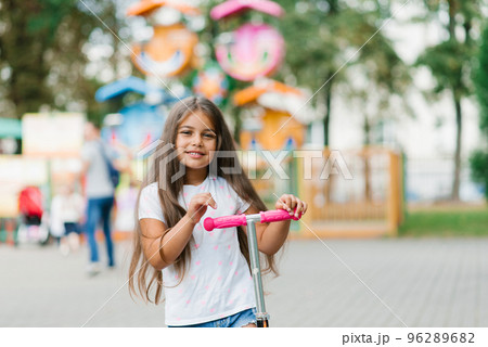 Beautiful smiling girl rides a scooter along the path of an amusement park on a summer day. Seasonal children's active sport. 96289682
