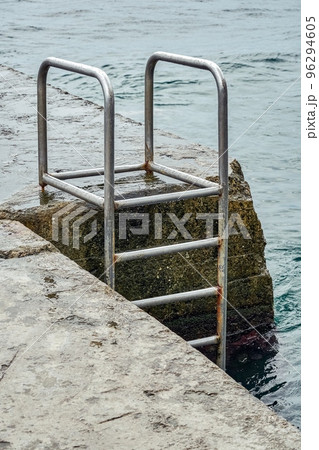 Grey concrete pier with metal ladder at blue sea water with small waves on an overcast summer day 96294605