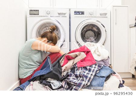 Upset woman in laudry room sitting ona floor with dirty clothes 96295430