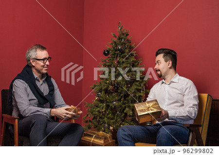 Elderly man and his adult son smiling and opening Christmas presents on red wall background 96299802
