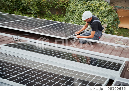 Man technician mounting photovoltaic solar moduls on roof of house. Builder in helmet installing solar panel system outdoors. Concept of alternative and renewable energy. Man technician mounting photovoltaic solar moduls on roof of house. Builder in helmet installing solar panel system outdoors. Concept of alternative and renewable energy. 96300656