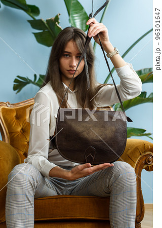 beautiful slender girl in gray checked pants and a white blouse posing while sitting in a yellow velor vintage armchair with a small black leather handbag beautiful slender girl in gray checked pants and a white blouse posing while sitting in a yellow velor vintage armchair with a small black leather handbag 96301647